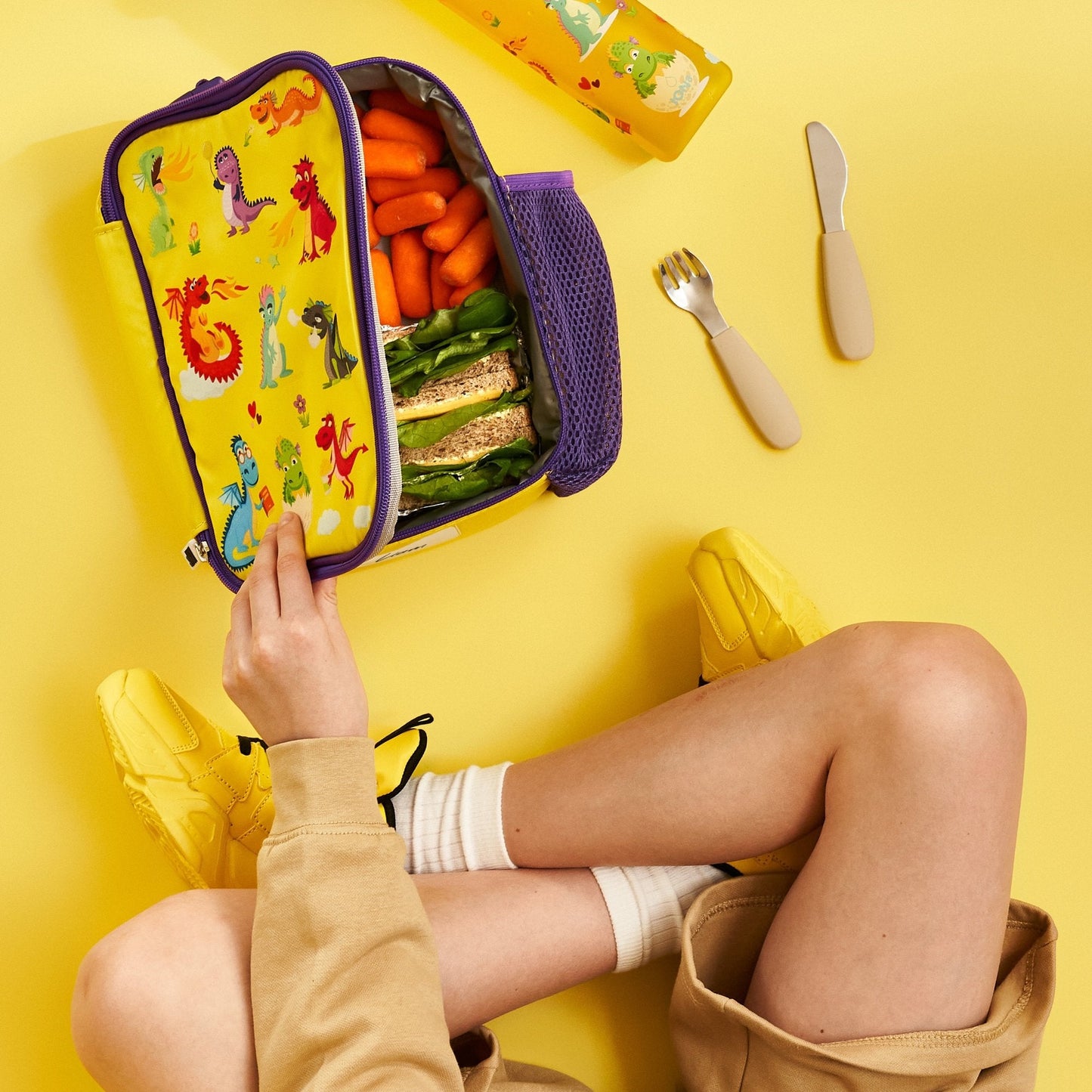 Person opening a colorful lunchbox with snacks on a yellow background