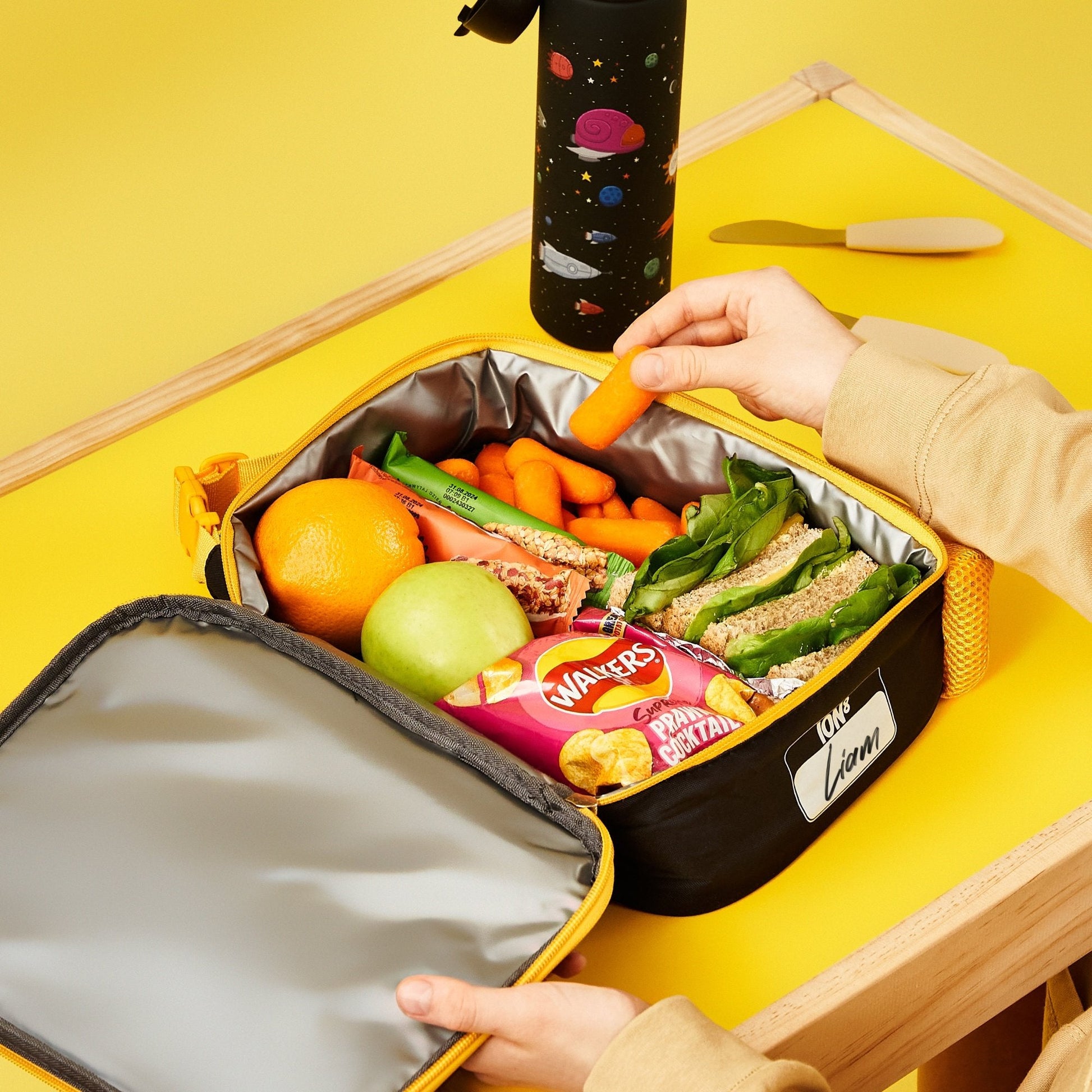 Lunch bag with food items including sandwiches, fruits, and a bottle on a yellow background