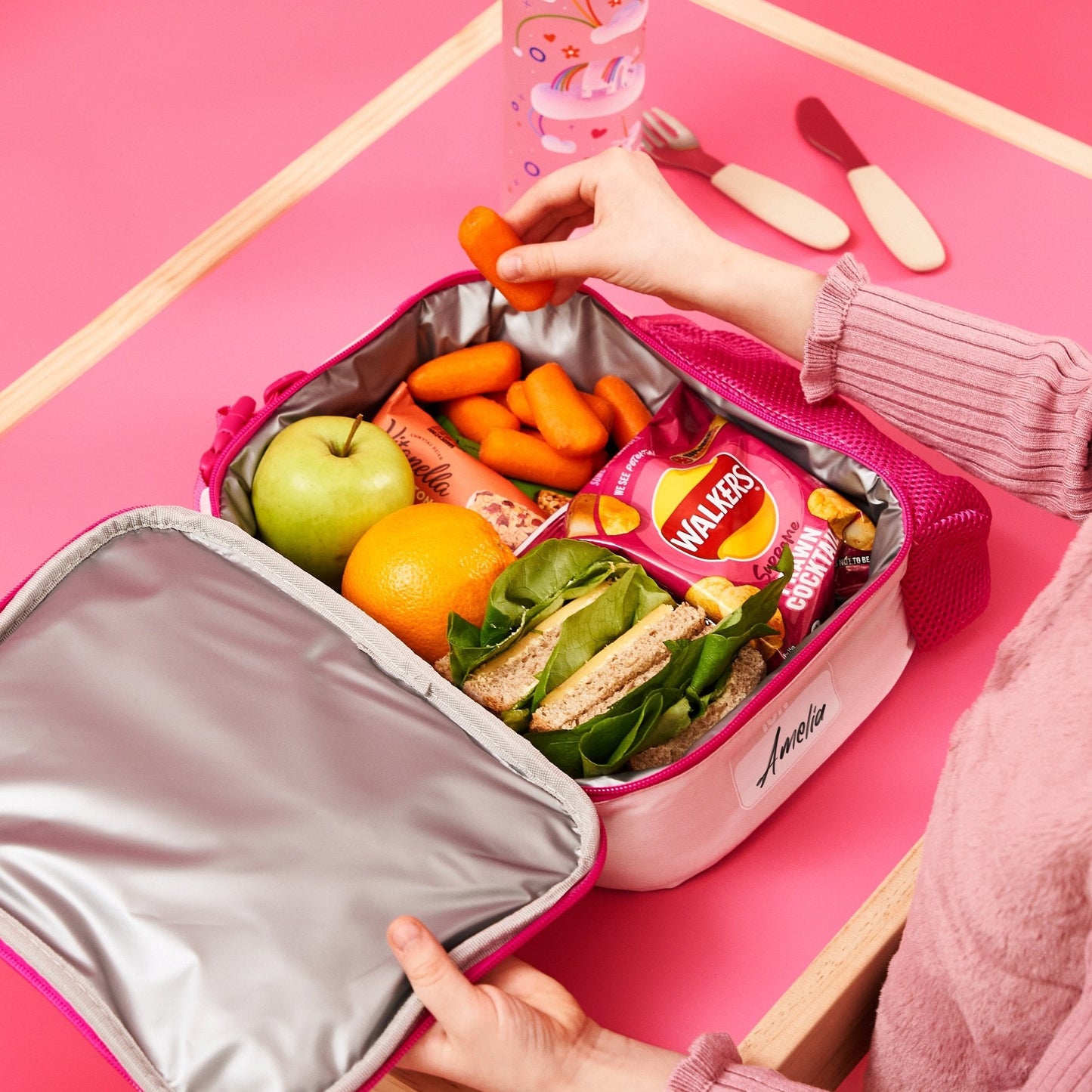 Person packing a lunchbox with fruits, vegetables, and snacks on a pink surface