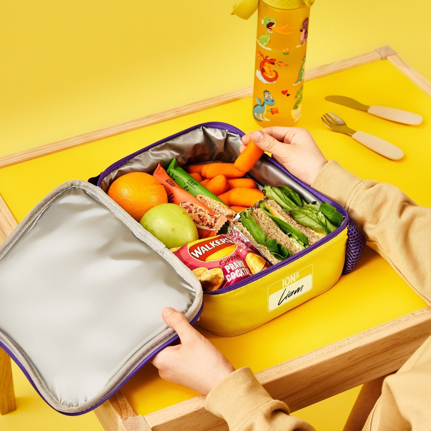 Yellow lunch box with a variety of food items on a yellow table with cutlery.