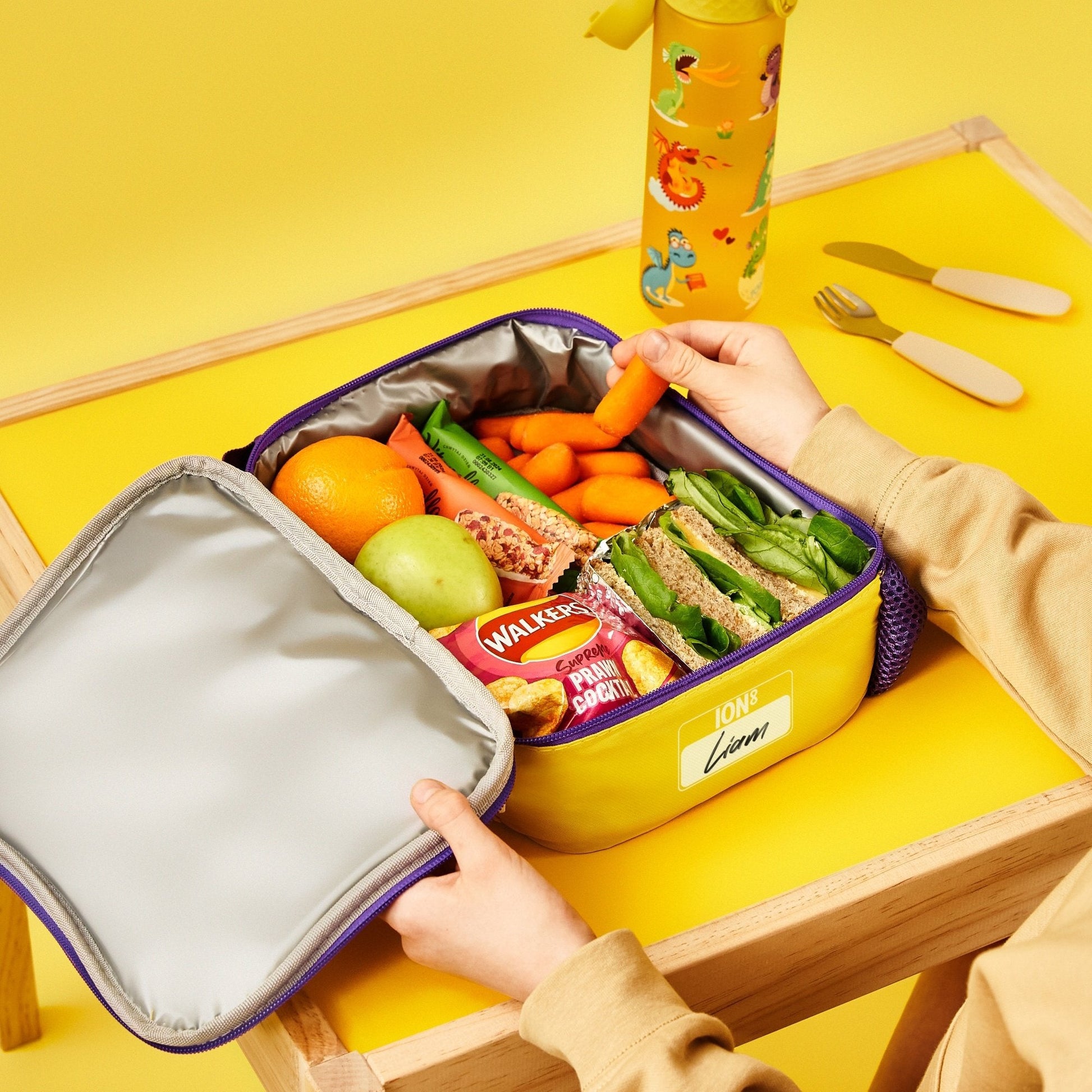 Yellow lunch box with a variety of food items on a yellow table with cutlery.