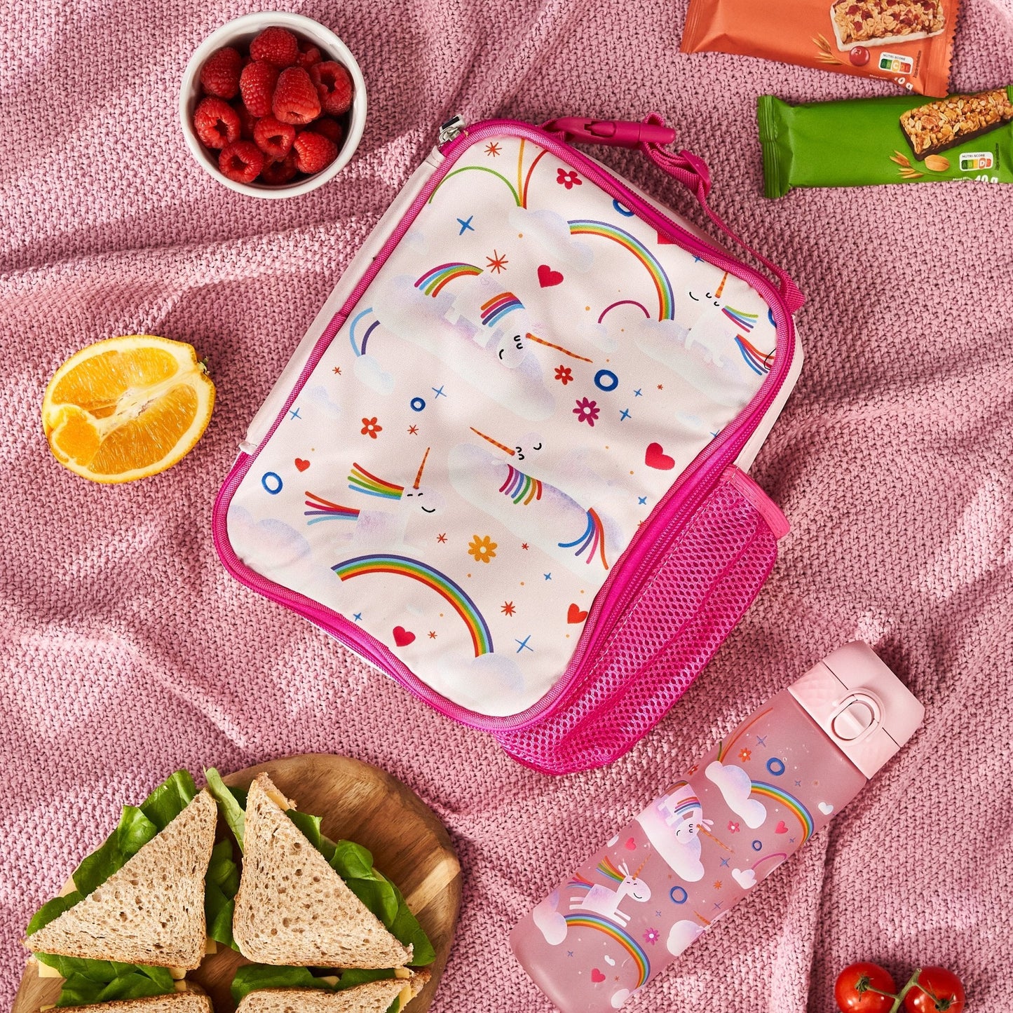 Lunch bag with unicorn and rainbow design on a pink blanket with snacks and sandwiches.