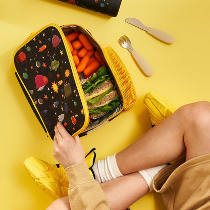 Person opening a lunch bag with a colorful pattern containing a sandwich and vegetables on a yellow background.