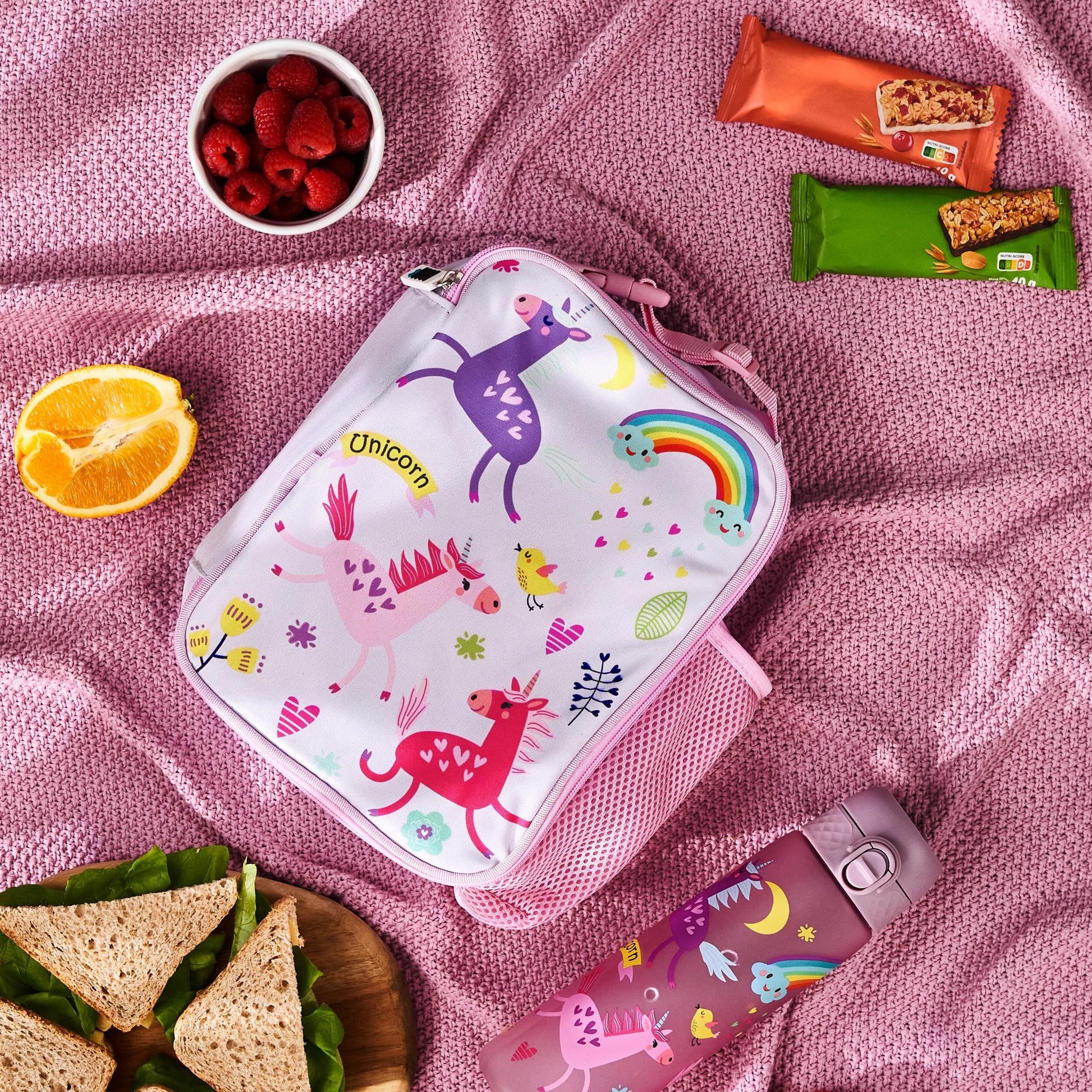 Pink lunch bag with unicorn design on a pink blanket with snacks and a water bottle.