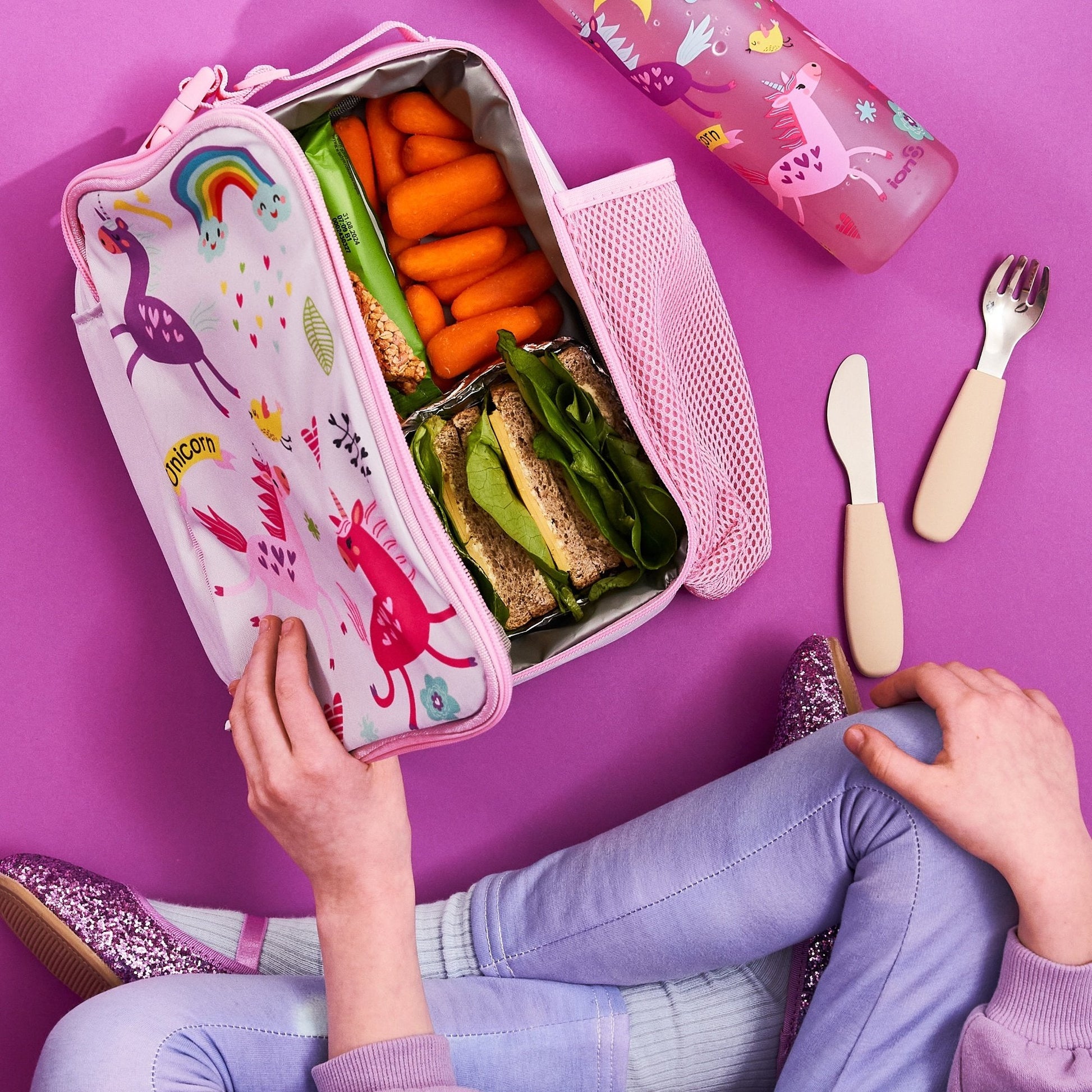 Pink lunch box with unicorn design filled with snacks on a purple background